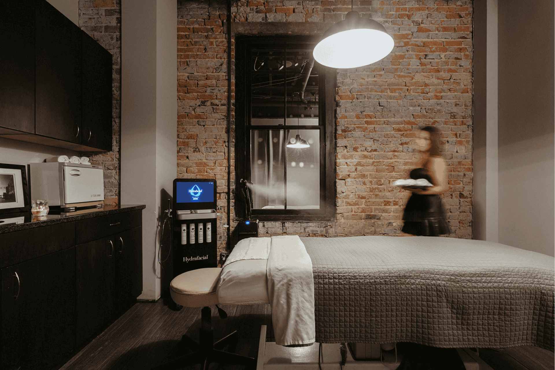 A woman carrying towels toward a bed by a Hydrafacial treatment machine