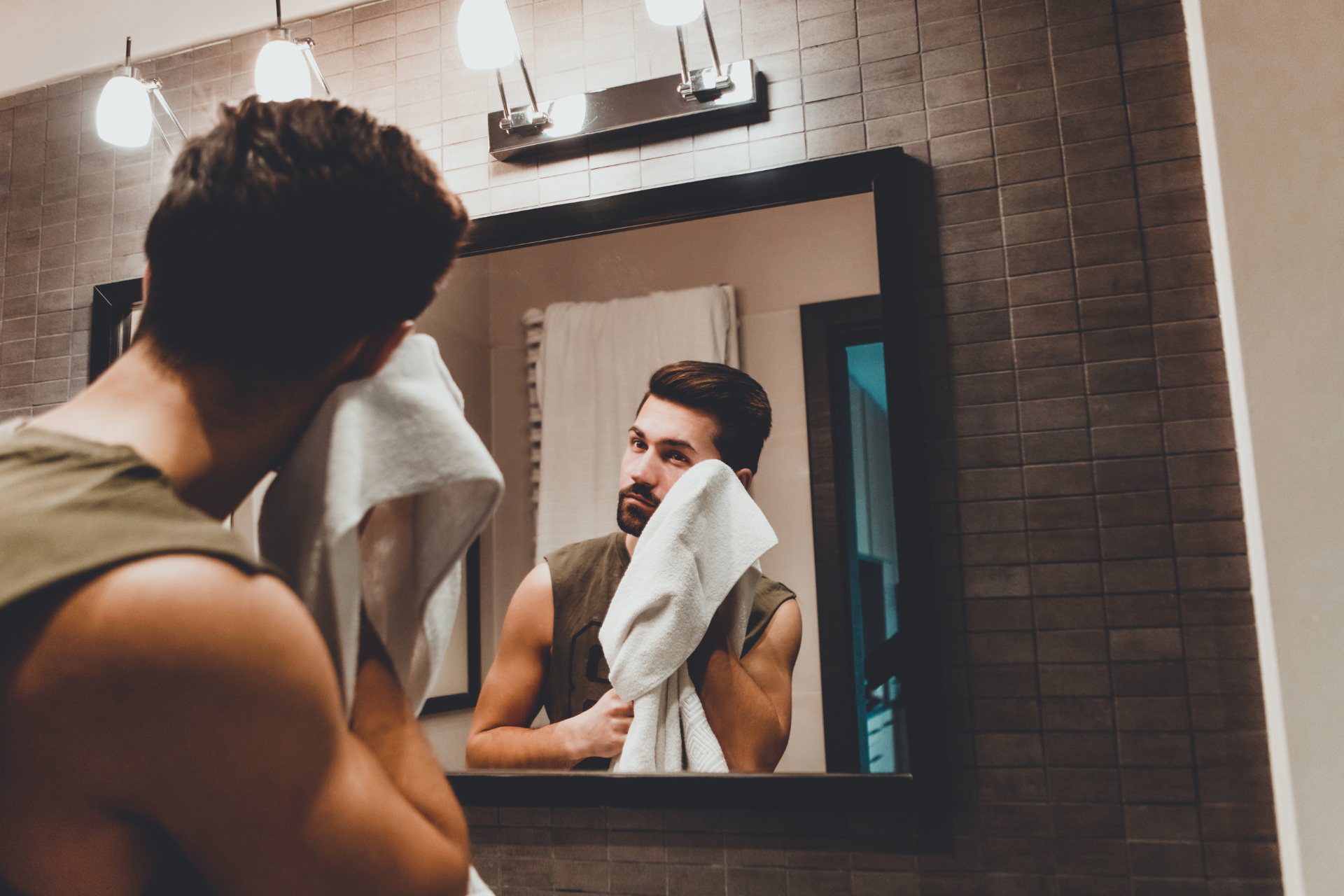 Young man patting his face with a towel while looking at his reflection in the mirror